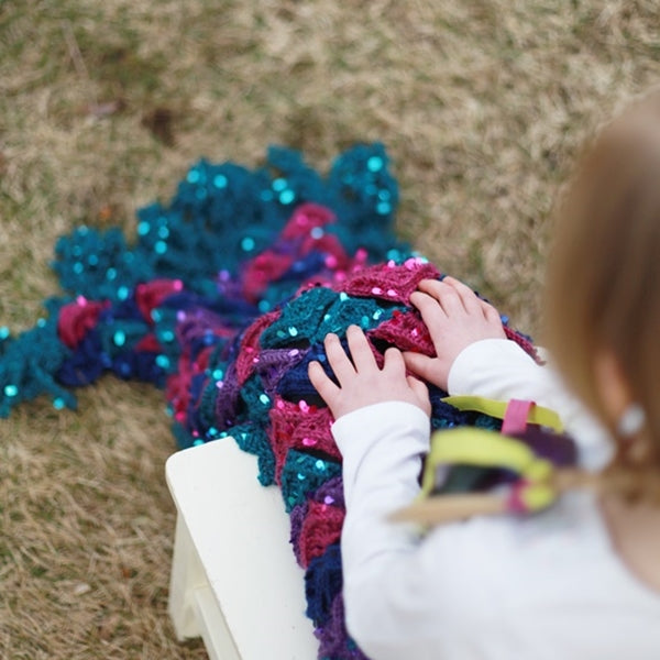 Little girl sitting with mermaid tail blanket in pink, purple, blue and teal, Mermaid Me Blanket, colorful crochet pattern by Susan Carlson of Felted Button