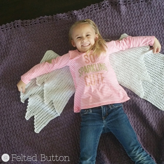 Cute little girl in pink shirt lying on purple blanket with white angel wings, smiling, Embraced By Angels Blanket crochet pattern by Susan Carlson | Felted Button | Colorful Crochet Patterns