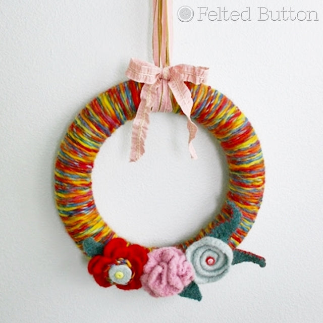 Wreath covered in rainbow yarn hanging by ribbon with felted flowers and buttons, Susan Carlson of Felted Button | Colorful Crochet Patterns
